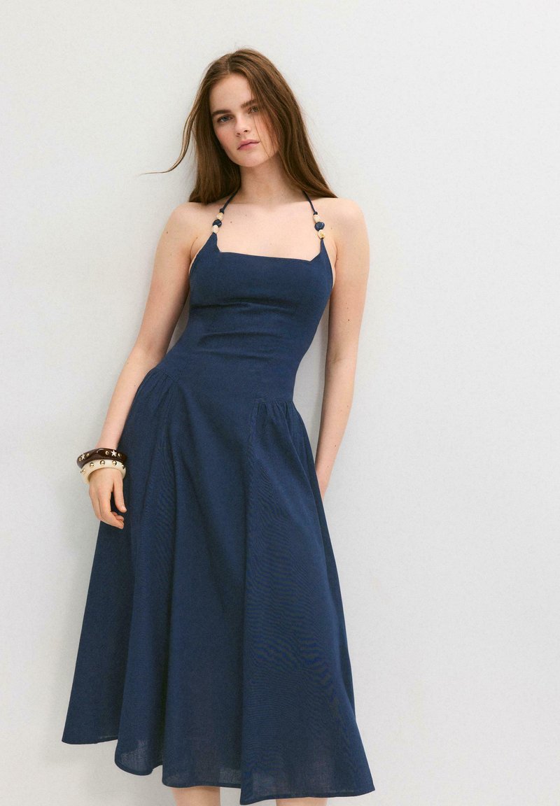 Jeune femme aux longs cheveux bruns portant une robe bleu marine à col licou et plusieurs bracelets, debout contre un mur blanc.