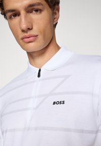 Hvit polo skjorte med glidelåskrage, med grå diagonale linjer og svart "BOSS"-logo på brystet. Glatt stofftekstur.