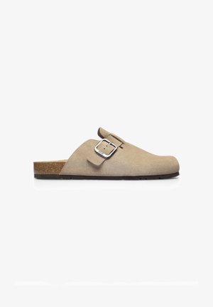 Zueco beige de ante sin cordones con hebilla plateada ajustable, entresuela de corcho y suela plana de caucho negro, visto de lado sobre fondo blanco.