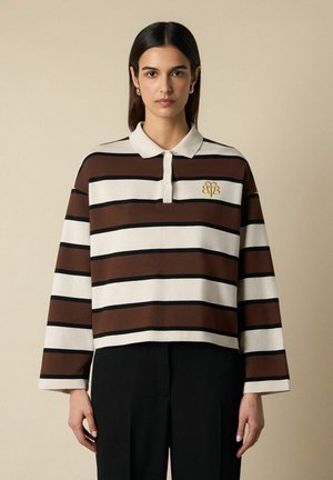 Vrouw met steil donker haar, gekleed in een bruin-wit gestreept poloshirt met lange mouwen en een zwarte broek, tegen een beige achtergrond.