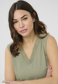 Haut sans manches vert olive avec un col en V, présentant des détails en dentelle élaborée autour de l'encolure. Texture lisse et coupe décontractée.
