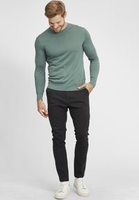 Lichtgroene, lange mouwen trui met een ronde hals, gecombineerd met zwarte slim-fit broek en witte sneakers op een neutrale achtergrond.
