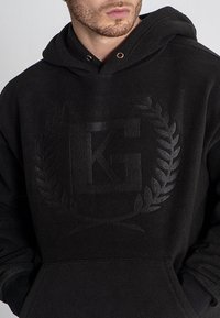 Sudadera de forro polar negra con un logotipo elevado rodeado de hojas de laurel, ojales dorados y un diseño clásico de bolsillo canguro. Textura suave.
