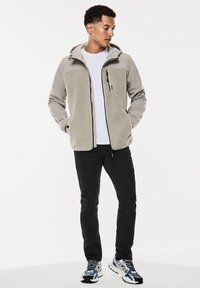 Ljusgrå fleecejacka med en slät tygdesign, framsida med dragkedja och huva. Åtföljs av svarta byxor och sneakers med blåa accenter.