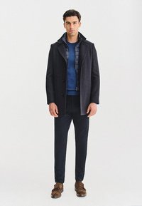 Ciemna kraciasta kołdra, niebieska ocieplana kurtka wewnętrzna, niebieska koszula, czarne spodnie i brązowe zamszowe buty. Gładka tekstura i dopasowany krój.