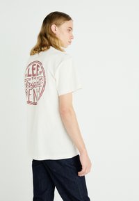 Cream-colored cotton t-shirt featuring a red round graphic print on the back. Short sleeves, relaxed fit, and hem hits at the hips.