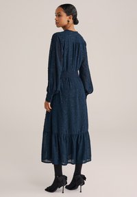 Marineblaues Langarmkleid aus strukturiertem Stoff mit gerafftem Taillenbereich und gestuftem Rock. Getragen mit schwarzen Strumpfhosen und Absätzen mit Schleifenakzenten.