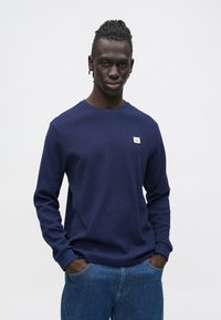 Tmavomodrá mikina s dlhými rukávmi a okrúhlym výstrihom, vyrobená z textúrovaného materiálu s malou logo nášivkou na hrudi. Kombinovaná s modrými džínami.