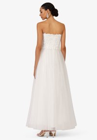 Robe blanche sans bretelles avec un corsage brodé, jupe en tulle fluide et fermeture éclair visible dans le dos. Le mannequin porte des sandales blanches à talons.