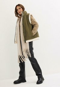 Gilet imbottito verde oliva con rifiniture in pelliccia sintetica bianca, abbinato a un maglione in maglia marrone, una sciarpa con frange crema e pantaloni e stivali in pelle nera.