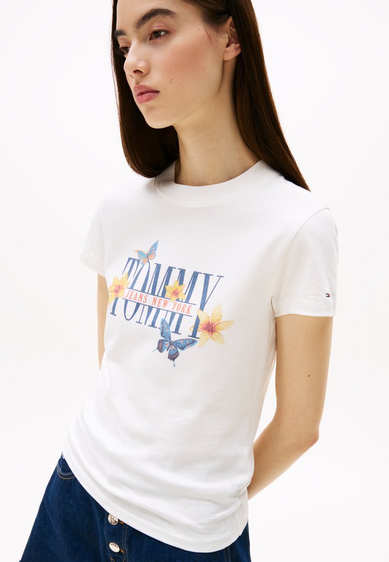 Camiseta blanca de algodón con gráficos de flores y mariposas. Cuello redondo y mangas cortas. "TOMMY JEANS NEW YORK" impreso en letras coloridas.