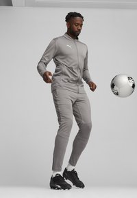 Grauer sportlicher Trainingsanzug mit einer Reißverschlussjacke und taillierten Hosen, aus strukturiertem Stoff mit einem Puma-Logo. Schwarze Fußballschnürer und ein Fußball.