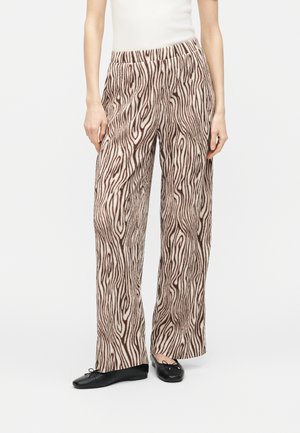 Kvinde iført vidde bukser med et brun- og beige træmønstret mønster, kombineret med sorte ballerinaer og en hvid ærmeløs top.