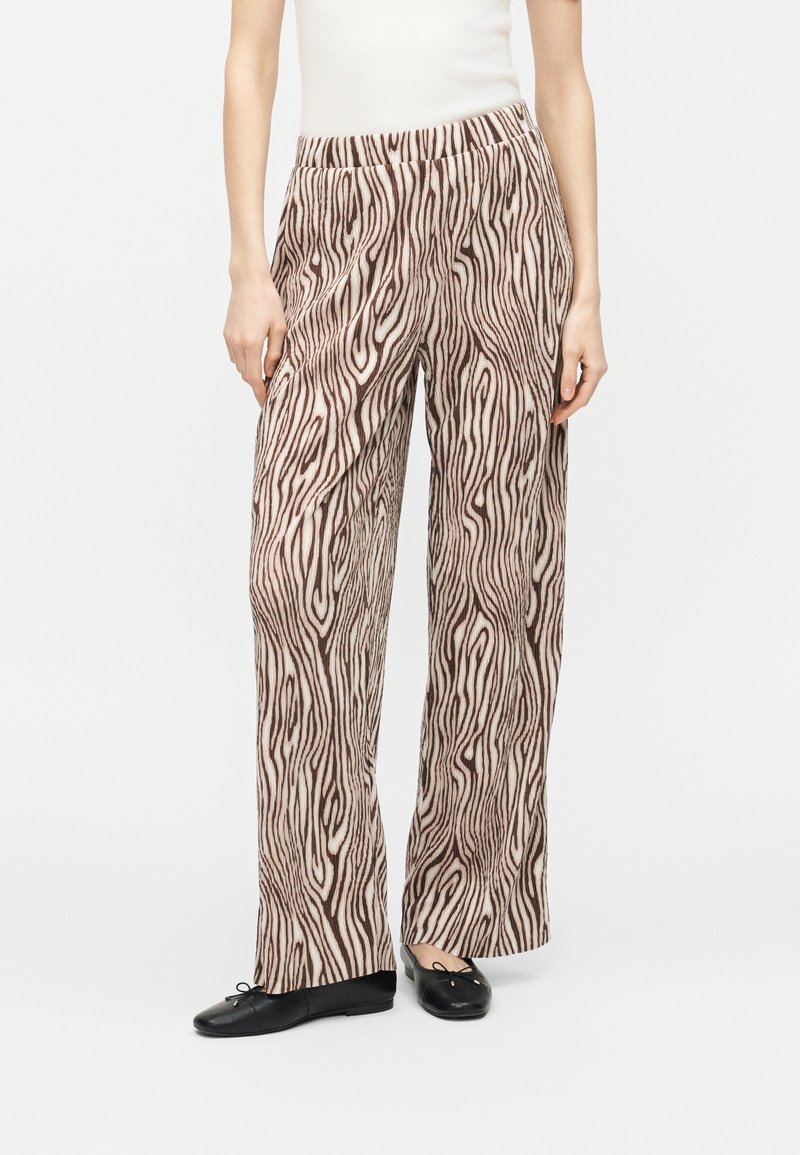 Γυναίκα που φοράει φαρδιά παντελόνια με καφέ και μπεζ σχέδιο ξύλου, συνδυασμένα με μαύρα μπαλέτα και λευκό αμάνικο τοπ.