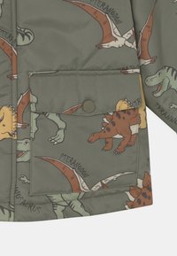 Tejido verde con dinosaurios impresos de colores, incluyendo Pteranodon, Tyrannosaurus y Stegosaurus, con un bolsillo que tiene un botón a presión.