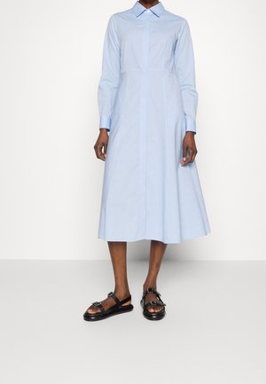 Kobieta w jasnoniebieskiej sukience midi z długim rękawem, kołnierzykiem oraz czarnych sandałach na odkryte palce z podwójnymi paskami i klamrami.