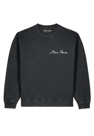 Junger Mann mit kurzen schwarzen Haaren, der einen schwarzen Pullover mit weißem Schriftzug „Mia Paris“ auf der Brust trägt und zur Seite schaut.