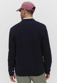 Mörkblå stickad cardigan med ribbad textur, som har krage och knäppta ärmslut, tillsammans med en ljusrosa basebollkeps.