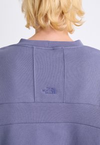 Paarse pullover met ribbelstructuur. Heeft een geborduurd logo in bijpassende kleur en een band over de achterkant. Standaard kraagontwerp.