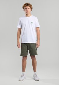 White short-sleeve t-shirt with a green palm tree graphic on the left chest, paired with olive green shorts and white sneakers.