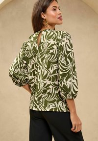 Bluse in grün-weißer Musterung mit Puffärmeln und einem Schlüsselloch-Rücken. Gewebter Stoff mit strukturierter Oberfläche. Kombiniert mit schwarzen Hosen.