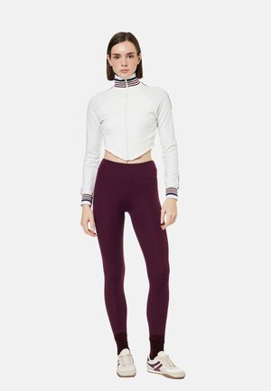Femme debout portant une veste courte blanche zippée avec col et poignets rayés, leggings violet foncé et baskets blanches avec des rayures.