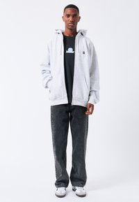 Felpa grigia con zip e tasca a marsupio, indossata sopra una maglietta nera. Abbinata a jeans grigio scuro e sneaker bianche. Design semplice.