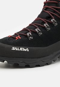 Melna pārgājienu zābaku ar teksturētu augšdaļu, sarkaniem šņorējumiem un metāla acīm. Tam ir izturīga zole, kas nodrošina saķeri, redzams logotips.