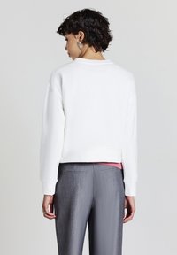 Felpa bianca corta con collo rotondo e maniche lunghe, abbinata a pantaloni grigi con trama e una maglietta rosa sotto. Design semplice.