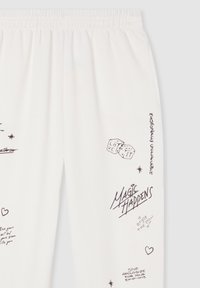 Białe spodnie z czarnym tekstem w stylu odręcznym i doodlami, w tym kostkami do gry, sercami, gwiazdami oraz frazami takimi jak "Magia się zdarza" i "Ciesz się chwilą".