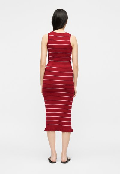 Γυναίκα που στέκεται με την πλάτη, φοράει αμάνικο κόκκινο φόρεμα με λευκές οριζόντιες ρίγες και βολάν στο τελείωμα, συνδυασμένο με μαύρα σανδάλια.