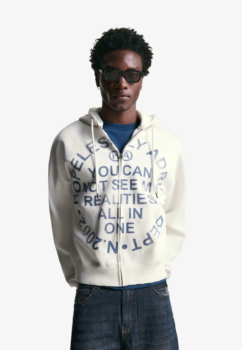 Cremefarbener Reißverschluss-Hoodie mit großen blauen Texten und Grafiken. Getragen über einem marineblauen Shirt, kombiniert mit dunklen Jeans. Quadratische Sonnenbrille.