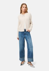 Kremowa kurtka boucle z zaokrąglonym kołnierzem i wiązaniem z przodu, zestawiona z szerokimi jeansami o prostych nogawkach, z kontrastowym przeszyciem i podwiniętymi mankietami.