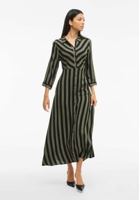 Dlhé košeľové šaty v olivových a čiernych vertikálnych pruhoch, s golierom a trojštvrťovými rukávmi, v kombinácii s čiernymi topánkami na podpätku.