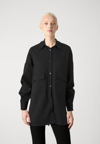 Giacca camicia nera a righe con maniche lunghe, chiusura con bottoni, due tasche sul petto e colletto a punta in tessuto leggero.