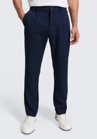 Pantalones azul marino con doble pinza, cierre de botón y bolsillos laterales, combinados con zapatillas blancas sobre un fondo liso.
