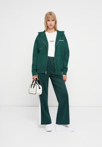 Felpa verde con zip e tasche frontali, abbinata a pantaloni larghi dello stesso colore e una t-shirt bianca. Gli accessori includono una piccola borsa bianca.
