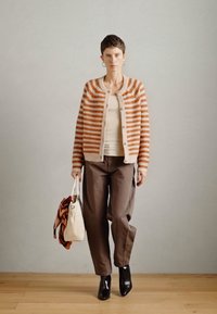 Cardigan a righe in arancione e beige, con chiusura a bottoni, abbinato a un top beige e pantaloni marroni larghi, con una borsa bianca.