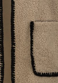 Beige strukturiertes Gewebe mit schwarzem Stich, ausgestattet mit einer aufgesetzten Tasche. Das Material hat ein weiches, flauschiges Aussehen mit kontrastierenden Details.