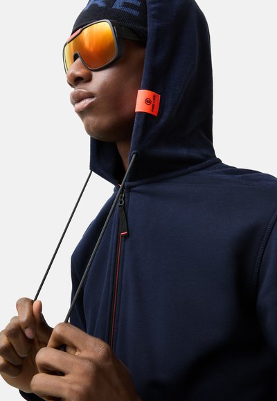 Navy hooded sweatshirt with a high collar, black zippers, and red accents. The person is wearing large orange-tinted sunglasses.