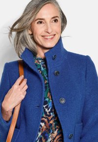 Königsblauer Wollmantel mit hohem Kragen, vier schwarzen Knöpfen und seitlichen Taschen, kombiniert mit einer farbenfrohen gemusterten Bluse.