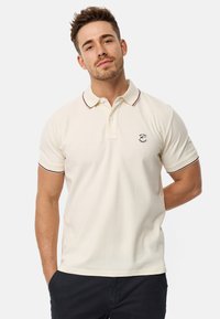 Beige Polohemd mit Kragen, ausgestattet mit bordauxrotem Besatz. Kurze Ärmel und ein kleines Logo auf der linken Brust. Glatte Stoffstruktur.