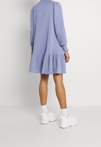 Vaaleansininen, pitkähihainen mekko, jossa on kerroksellinen röyhelöhelma; valmistettu tekstuuri-kankaasta, yhdistettynä valkoisiin paksupohjaisiin tennareihin ja matalapohjaisiin sukkiin.