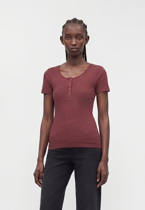 Femme aux cheveux tressés portant un haut bordeaux côtelé ajusté à manches courtes avec quatre boutons, associé à un pantalon noir sur fond blanc uni.