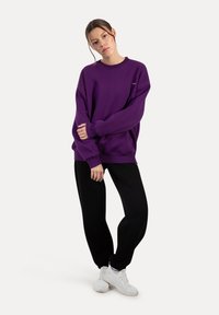 Sudadera morada de manga larga, con corte relajado, puños de canalé y cuello redondo, combinada con pantalones negros y zapatillas blancas.