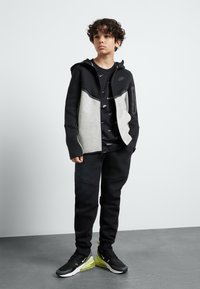 Nike dukserica na zip u crnoj i svijetlo sivoj boji, uparena s crnim trenirkama i crno-zelenim tenisicama. Sadrži kapuljaču i više džepova.