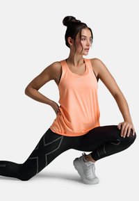 Leichtes oranges Tanktop mit einem strukturierten Muster, kombiniert mit schwarzen Leggings, die ein graues, kantiges Design aufweisen. Weiße Sportschuhe.