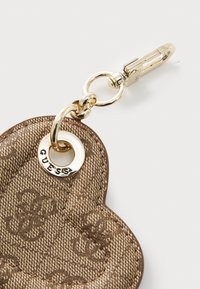 Breloc de cheie imprimat cu logo de culoare maro, în formă de inimă, având un clips din metal de nuanță aurie și un inel circular cu inscripția "GUESS".