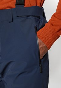 Námořnické voděodolné kalhoty mají zipovou kapsu, nastavitelné pas a texturovanou tkaninu. Oranžový pletený svetr je viditelný vedle zkráceného obrázku.