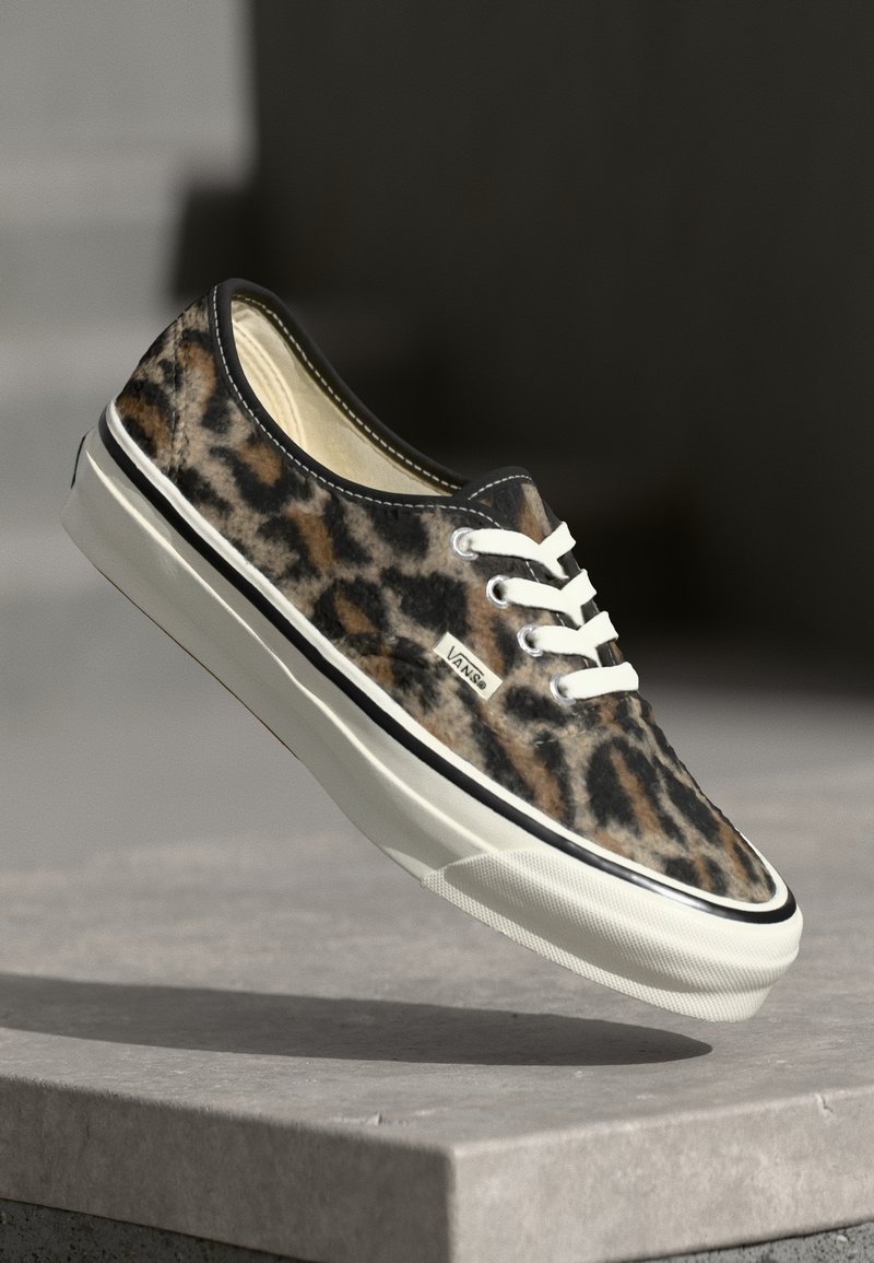 Baskets Vans à imprimé léopard avec lacets et semelle blancs, légèrement surélevées au-dessus d'une surface en pierre grise sur un fond sombre et flou.